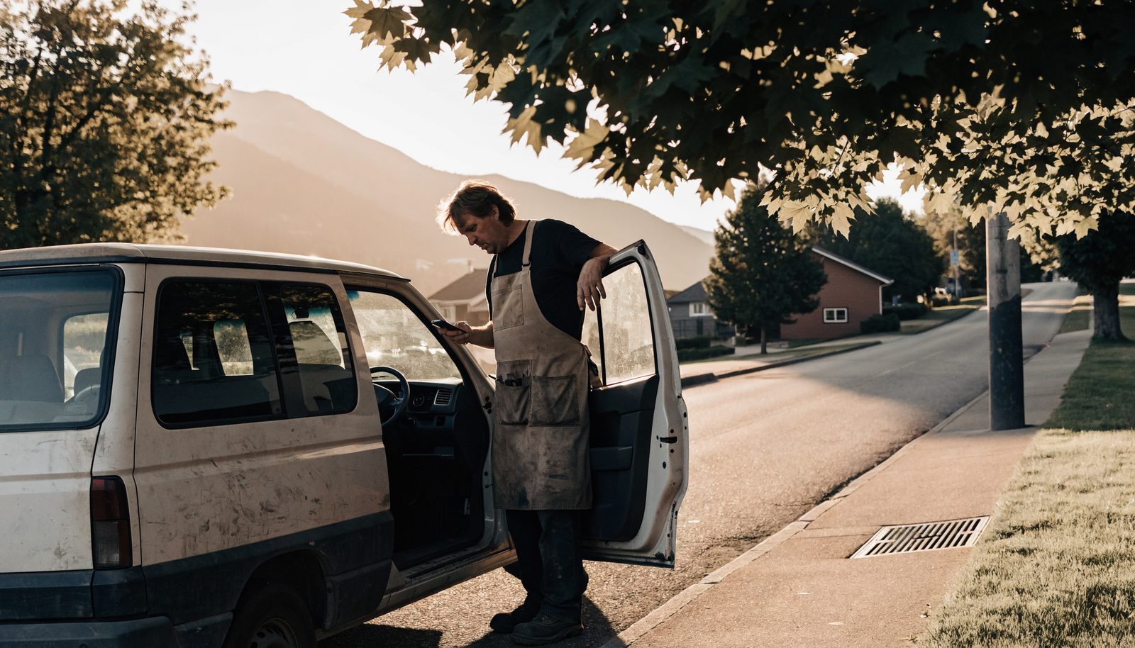 A Canadian tradesperson at a work van, checking a phone on a quiet street.